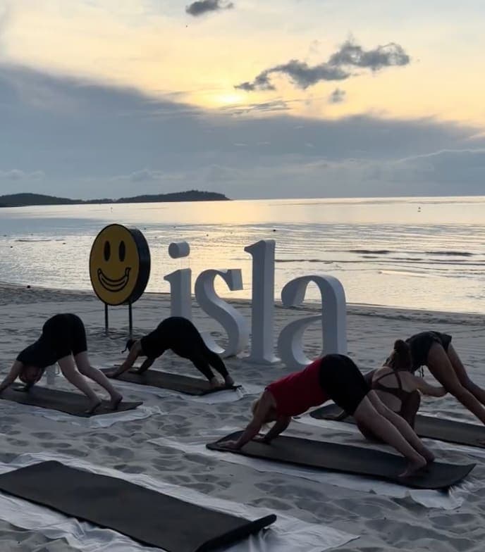 A yoga class in downward dog on the sand at sunrise in Koh Samui, beside the studio's letters