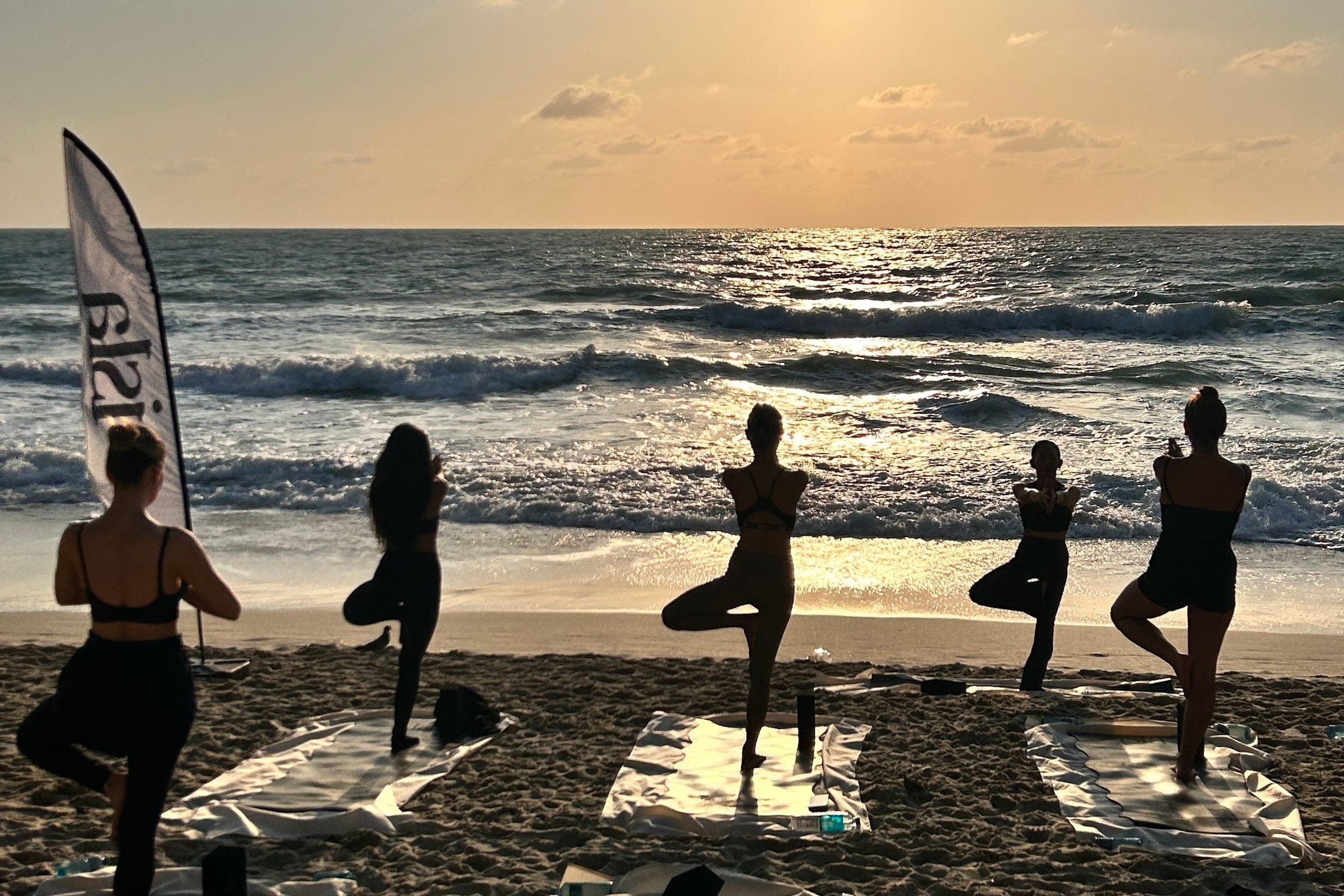 Five people in tree pose on mats on the sand at sunrise, the Isla flag beside them — the beachfront yoga class at Banana Fan Sea Resort, Koh Samui